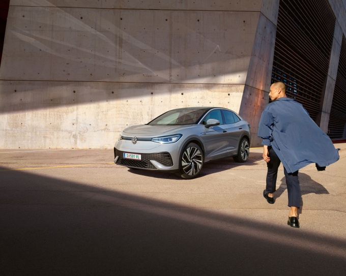 Ein silberner VW.ID 5 (Elektroauto) in modernem Design, geparkt vor einer Betonwand in urbanem Ambiente. Im Vordergrund geht ein Mann in einem blauen Mantel auf das Fahrzeug zu.