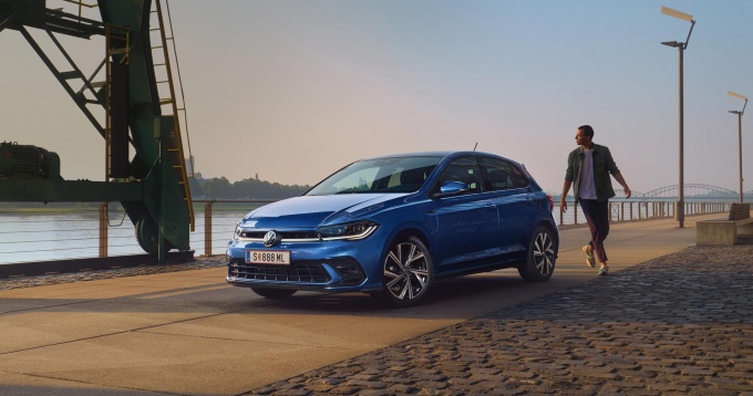 Ein blauer VW Polo R-Line geparkt auf einer gepflasterten Uferpromenade mit Blick auf einen Fluss bei Sonnenuntergang. Eine Person im legeren Outfit läuft neben dem Fahrzeug entlang.