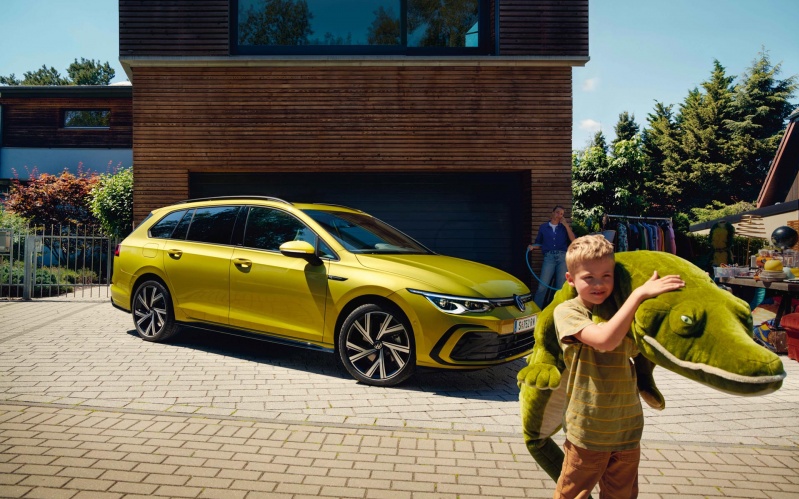 Ein gelb-grüner Golf Variant R-Line parkt vor einem modernen Wohnhaus mit Holzverkleidung und großen Fenstern. Im Vordergrund hält ein fröhliches Kind ein großes Plüschtier in Form eines grünen Krokodils.