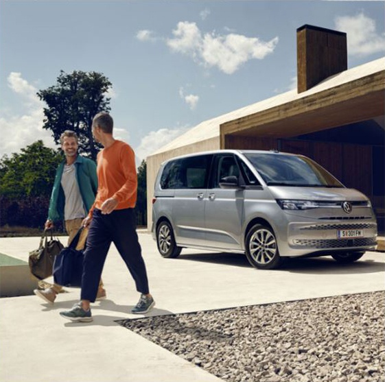 Ein silberner VW Multivan parkt auf einer modernen Auffahrt vor einem Haus mit klaren, minimalistischen Linien. Im Vordergrund unterhalten sich zwei Männer lächelnd, einer trägt eine orangefarbene Jacke, der andere eine grüne, beide halten Taschen in den Händen.