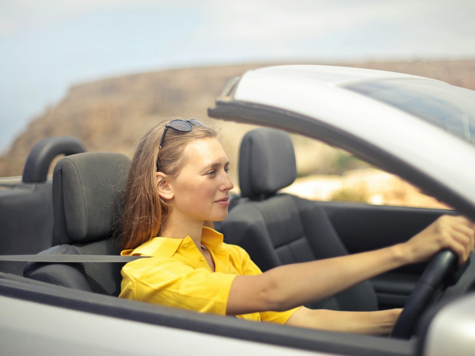 Eine junge Frau in einem Sommer-Outfit fahrt ein Cabrio mit offenem Verdeck.