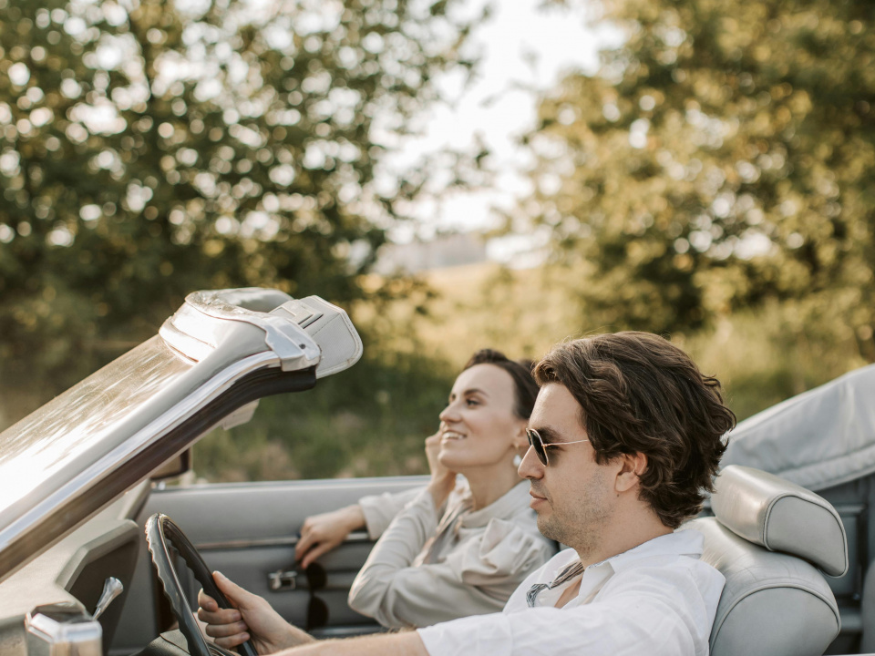 Ein Paar f&auml;hrt in einem Cabrio umgeben von bl&uuml;henden B&auml;umen.