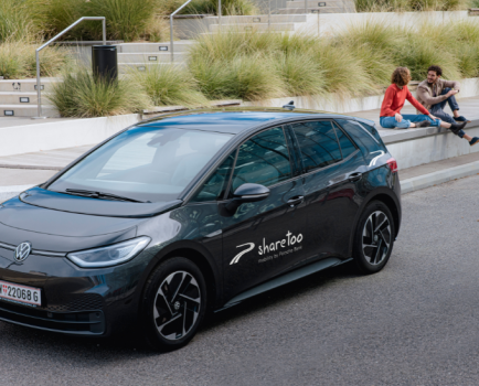 dunkles parkendes Elektroauto mit der Aufschrift sharetoo mit 2 Erwachsenen Menschen im Hintergrund