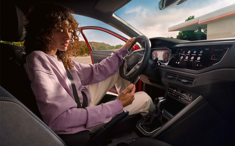 Eine Frau mit lockigem Haar sitzt in einem Volkswagen mit geöffnetem Fahrerfenster und geöffneter Tür. Sie trägt einen lila Pullover und hält ein Smartphone in der Hand, während sie die andere Hand am Lenkrad hat. Das moderne Armaturenbrett zeigt ein digitales Cockpit und ein großes Infotainment-Display. Im Hintergrund ist eine Tankstelle und Natur zu sehen.