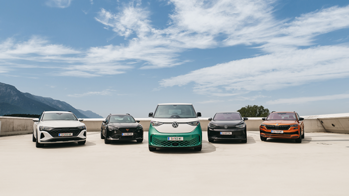 Ein Audi A5, ein Cupra Ateca, ein VW ID.Buzz und ein VW ID.5 sowie ein Skoda Karoq in verschiedenen Farben stehen nebeneinander auf einer offenen Fläche. Im Hintergrund sind Berge, Bäume und ein strahlend blauer Himmel mit weißen Wolken zu sehen.