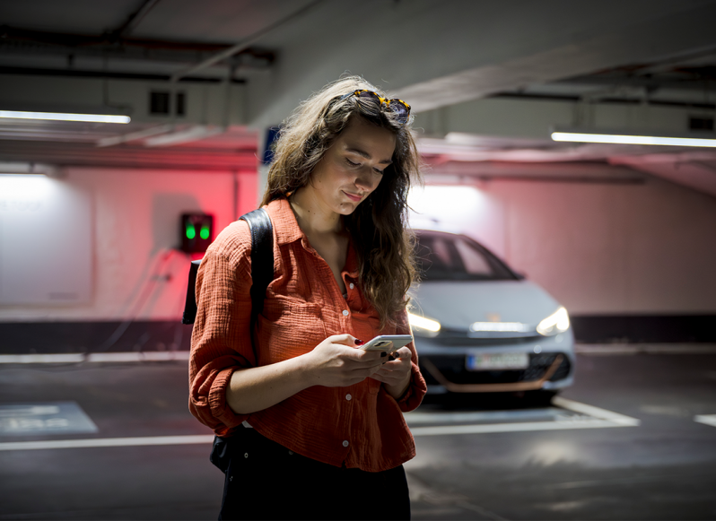Frau mit roter Bluse schaut auf Handy vor parkendem Auto in einer Tiefgarage