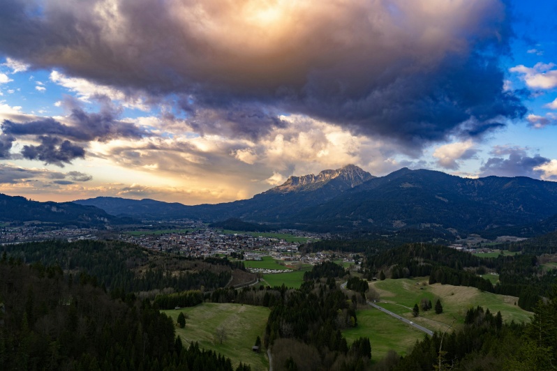 Blick auf die Berge und das Tal von einem Hügel aus, mit grünen Wiesen im Vordergrund und blauen Himmel darüber.